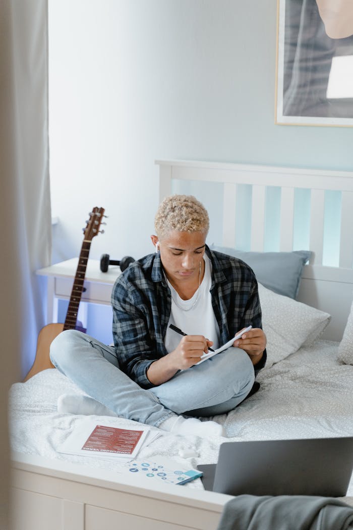 Young man studying in bedroom with laptop and notebook, embodying online learning.