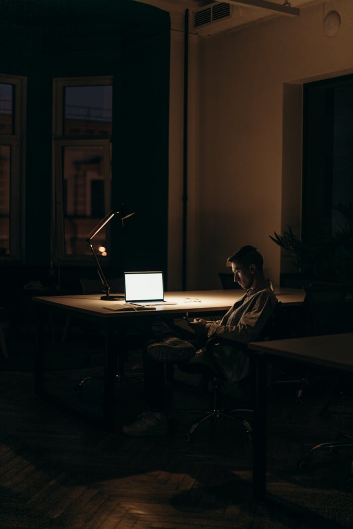 A lone man working on a laptop under dim lighting in a quiet office at night.